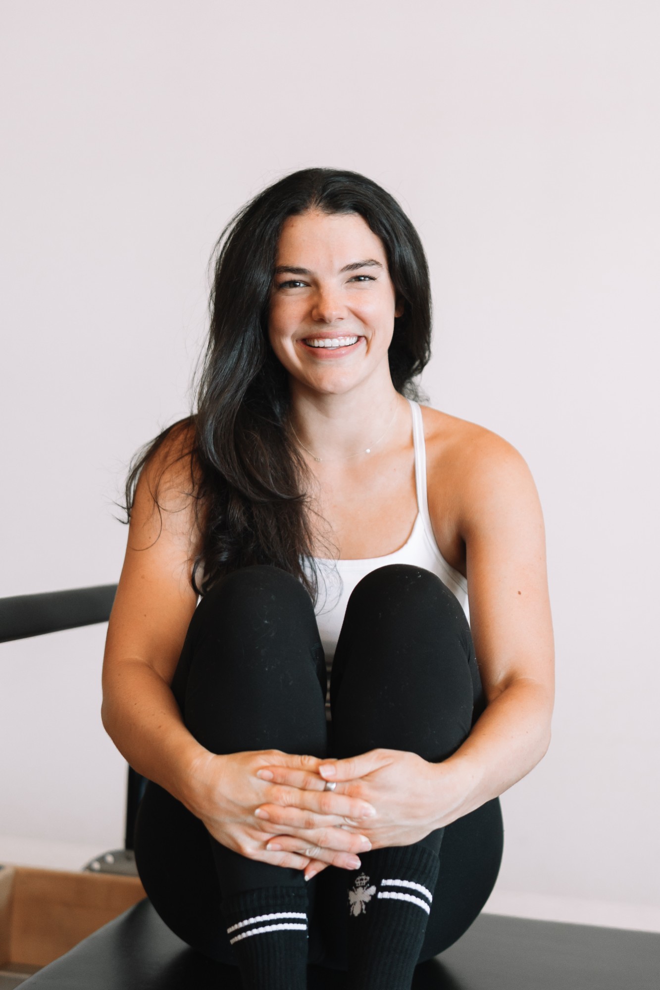 Brett smiling, sitting on a reformer hugging her knees